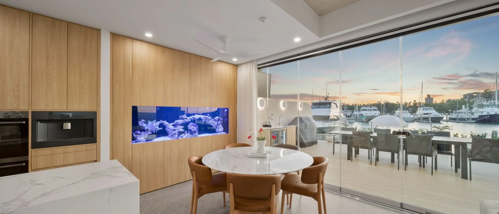 Kitchen and dining area of a completed waterfront home, timber joinery, marble island and glazing opening onto the terrace at dusk