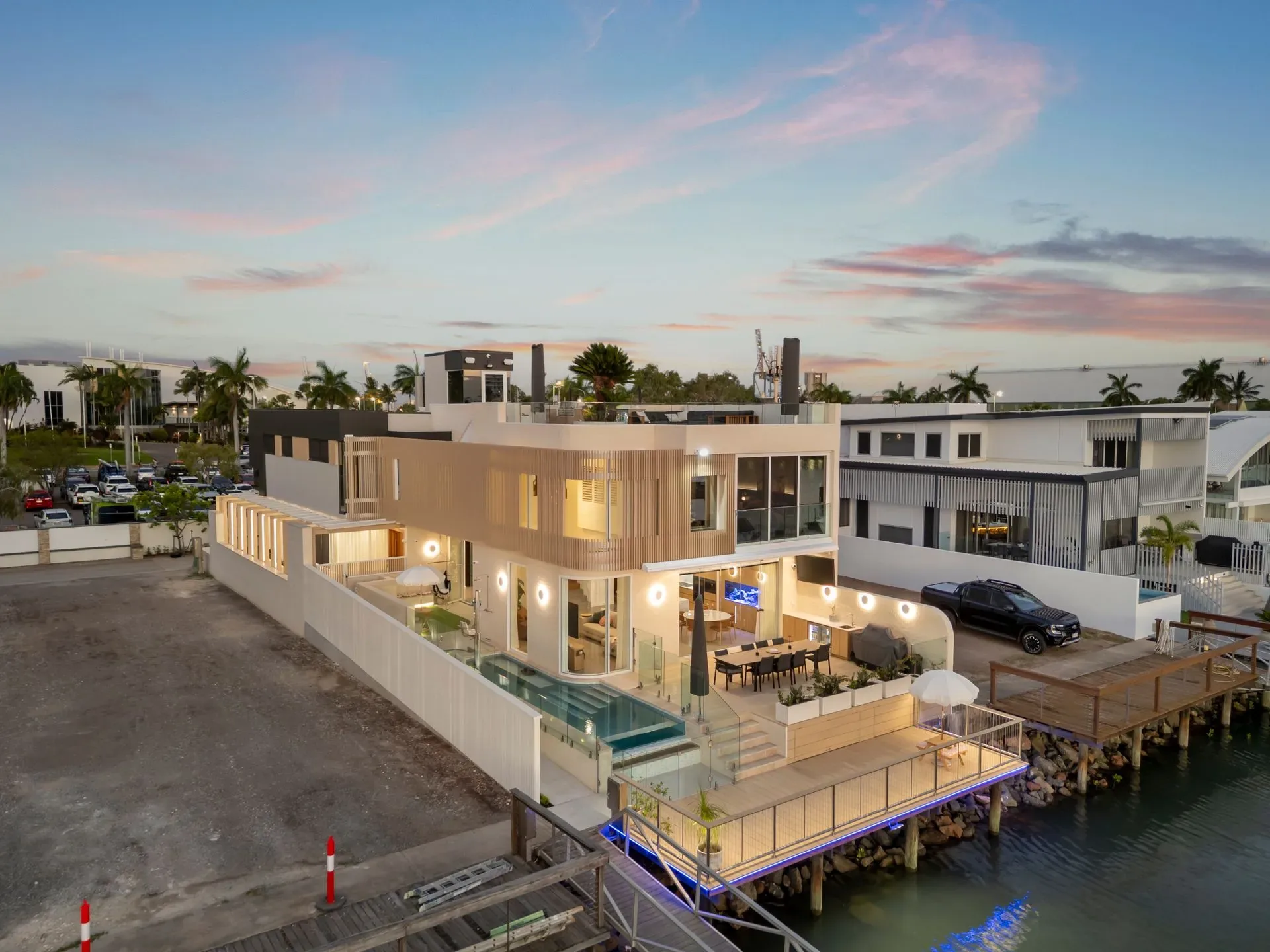 Completed waterfront home at dusk seen from across the water, pool and terraces lit
